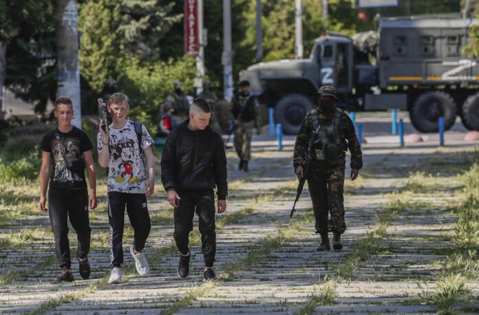 Nuotraukoje, darytoje per Rusijos kariuomenės organizuotą ekskursiją žiniasklaidai, matyti, kaip Skadovske, Chersono srityje, Ukrainoje, vietiniai jaunuoliai eina pro Rusijos karį.