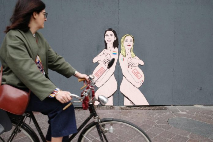 Moteris šalia Italijos ministrės pirmininkės Giorgios Meloni (dešinėje) ir PD lyderės Elly Schlein pavaizdavimo, kurį nupiešė menininkas aleXsandro Palombo Milane, Italijoje / EPA nuotr.